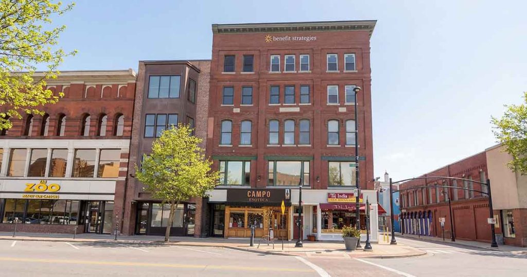 A street view of a multi-story red brick building with businesses on the ground floor, including Campo Enoteca restaurant and The Daily Scoop, with trees and a clear blue sky.