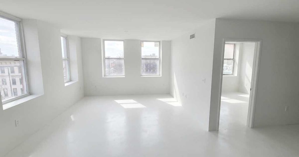 A bright, empty apartment room with white walls and floors, large windows letting in natural light, and an adjoining smaller room visible through an open doorway.