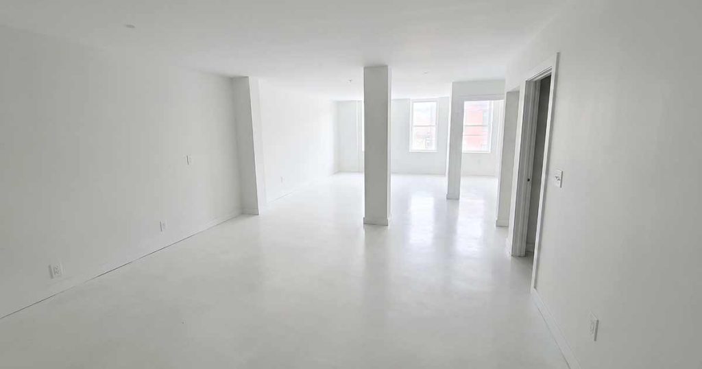 A spacious, empty white room with two central pillars, large windows letting in natural light, and an open doorway on the right side. The walls, floor, and ceiling are all painted white.