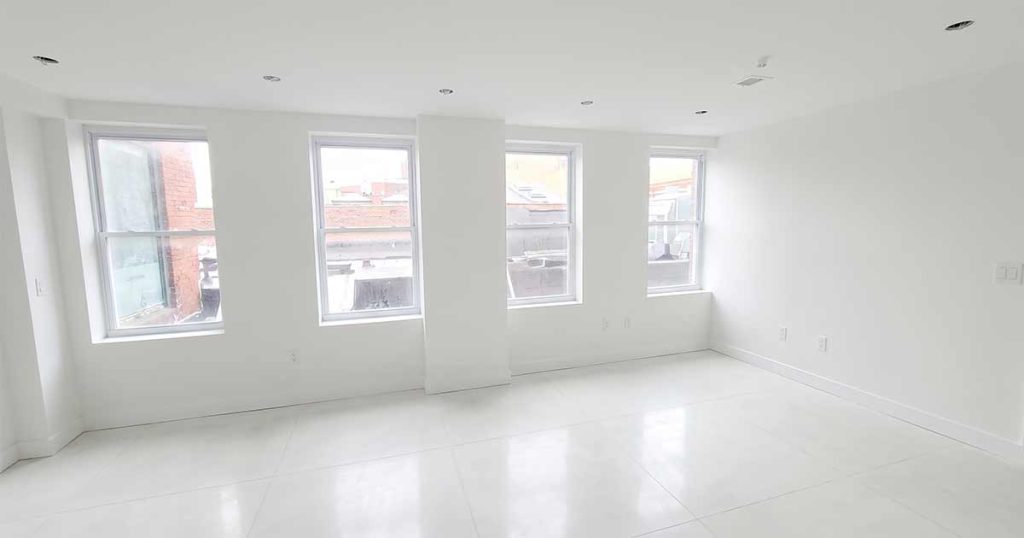 A bright, empty room with large white tile floors and four tall windows letting in natural light, with white walls and ceiling. Urban buildings are visible through the windows.