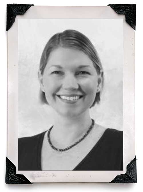 Black-and-white portrait of a smiling woman with short hair, wearing a dark top and a beaded necklace, displayed in a photo frame with black corner protectors.