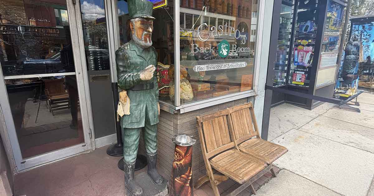 A wooden statue of a bearded man in a green uniform stands outside a storefront near two wooden chairs and a trash can. The window displays signs for a back room and a craft beer establishment.