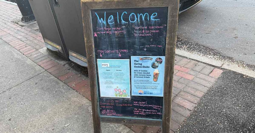 A chalkboard sidewalk sign welcomes visitors, displaying handwritten chalk messages and posters about local events and Girl Scout Cookie ice cream sandwiches, positioned on a sidewalk near the street.