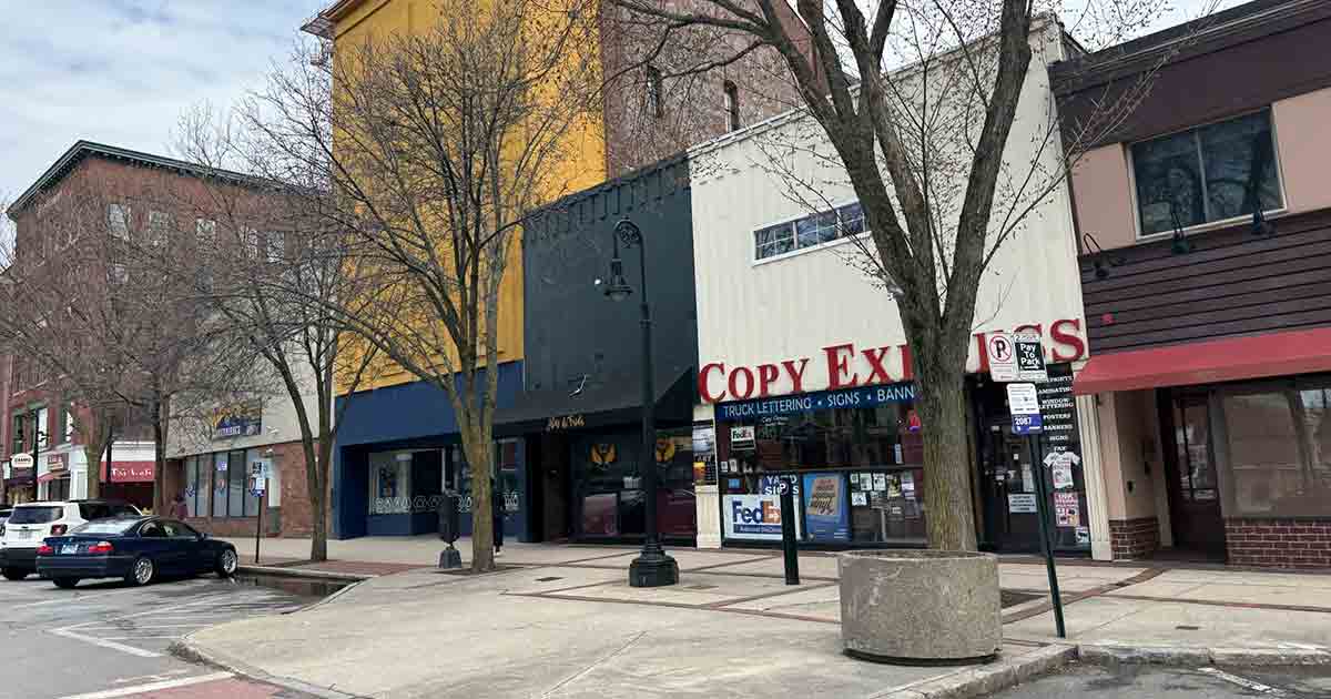 A row of small businesses on a city street, including a print shop called Copy Express. Leafless trees line the sidewalk, and a few cars are parked along the curb under a cloudy sky.
