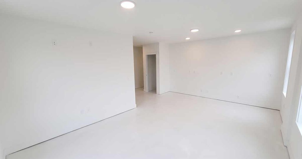 Empty, unfurnished room with white walls, white floor, and recessed ceiling lights. There are no decorations or furniture, and a doorway opens to another space. Large windows on the right let in natural light.
