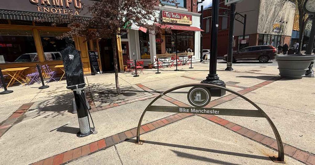 A downtown sidewalk in Manchester with a “Bike Manchester” rack, outdoor restaurant seating, a parking meter, and shops including Campo Enoteca and Cafe Mo India. A tree and parked cars are visible.