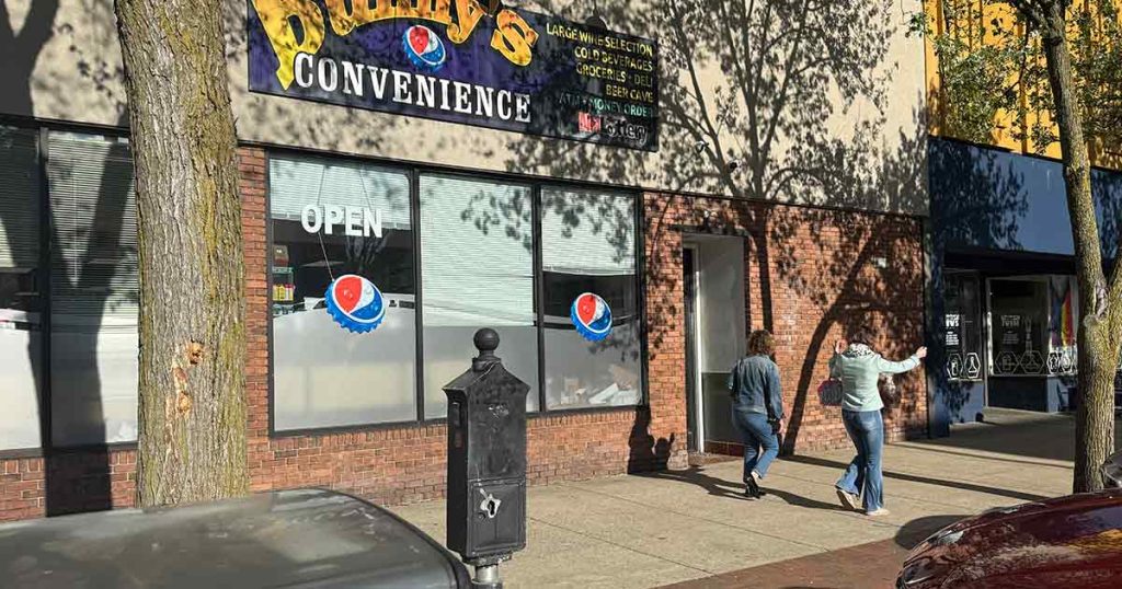 Two people walk on a sunny sidewalk past a store called "Duffy's Convenience," which has an "OPEN" sign and window decals. Trees cast shadows on the storefront and sidewalk.