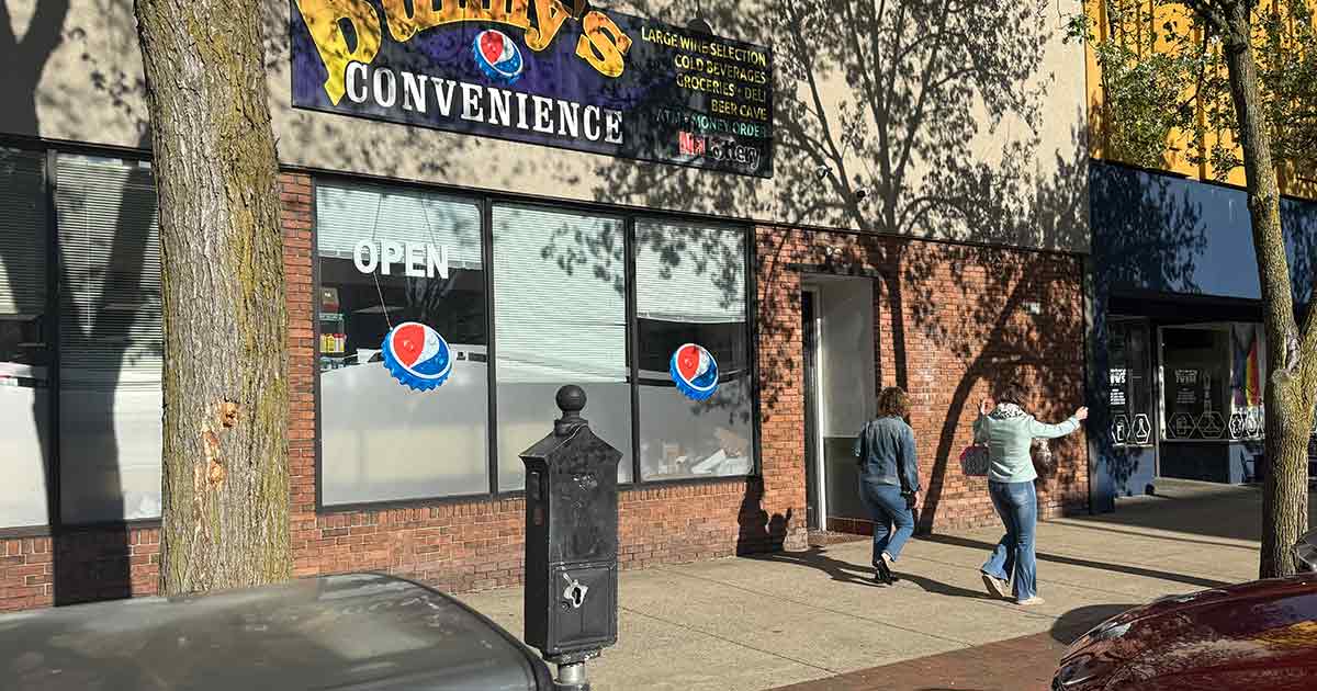 Two people walk on a sunny sidewalk past a store called "Duffy's Convenience," which has an "OPEN" sign and window decals. Trees cast shadows on the storefront and sidewalk.