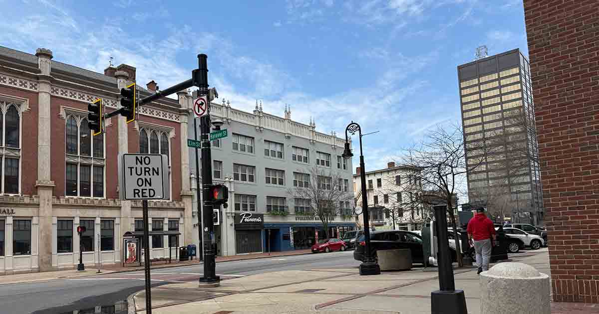 A city street corner with traffic lights, a "No Turn On Red" sign, and various buildings in the background. A person in a red jacket is walking on the sidewalk. The sky is partly cloudy.
