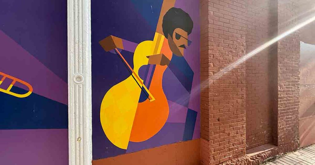 Colorful mural of a man with sunglasses and a mustache playing a yellow-orange cello on a purple background, with sunlight streaming across a red brick wall.