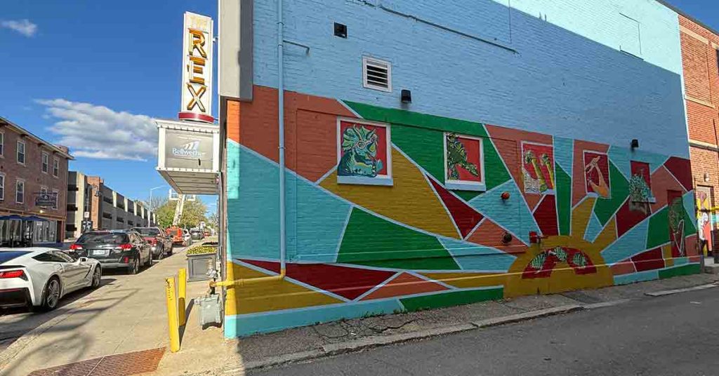 A colorful mural with geometric shapes and framed images decorates the side of a building next to the REX Theatre sign. Cars are parked along the street under a blue sky with scattered clouds.
