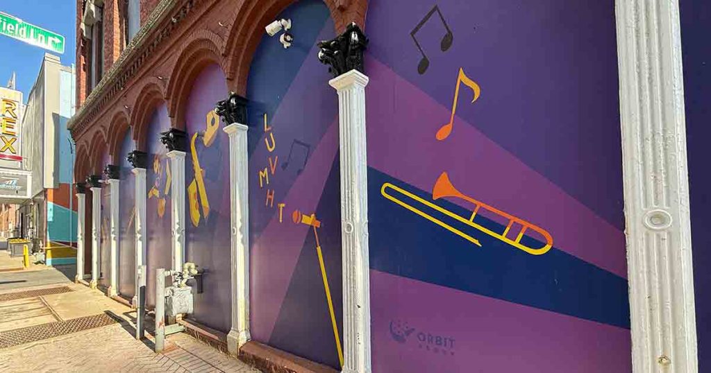 A purple mural on a building features yellow musical notes, a saxophone, trumpet, and trombone, with white columns in front. Nearby signs read "REX" and "Field Inn.