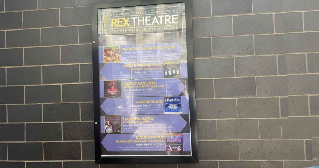 A poster for the Rex Theatre displays upcoming event listings with dates and colorful graphics, mounted on a dark brick wall. The headings and event details are visible but not fully readable.