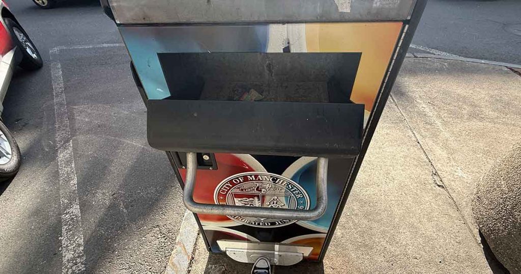 A colorful outdoor parking meter with a City of Manchester sticker on the front stands on a sidewalk next to a street. The meter is partially in the sun and has a shadow cast over its screen.