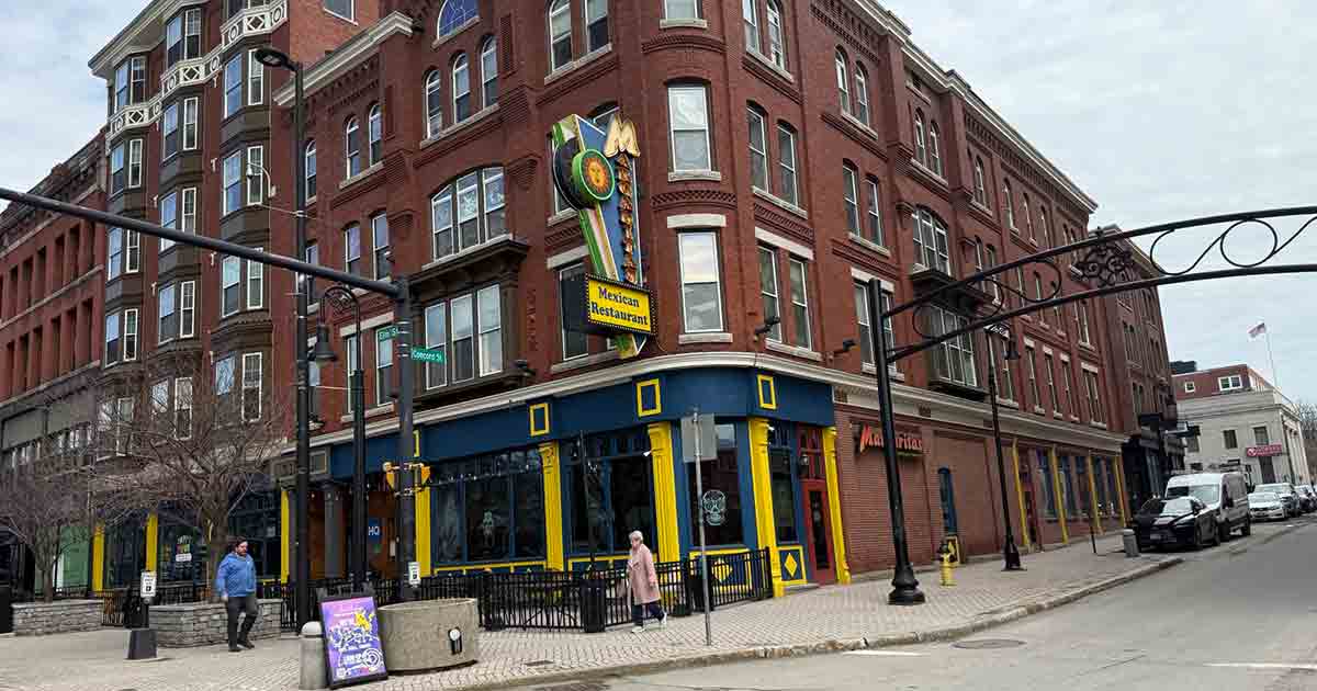 A street corner in a city with a red brick building housing a restaurant called "Victoria Restaurant." Pedestrians walk on the sidewalk, and cars are parked along the street under a metal archway.