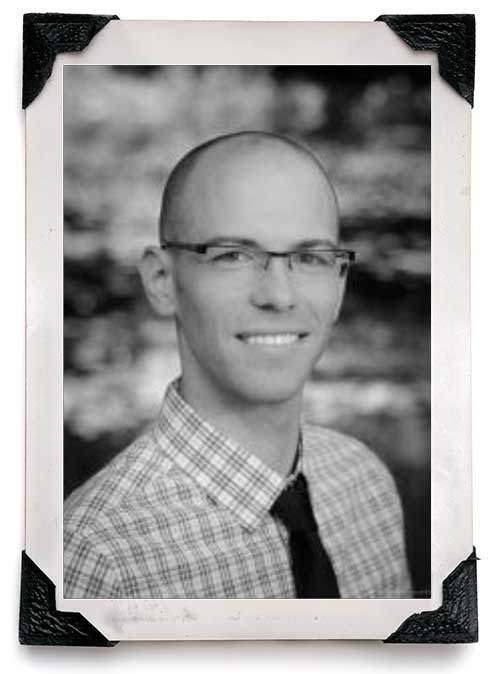 Black-and-white portrait of a smiling, bald man wearing glasses, a plaid shirt, and a dark tie. The photo is framed with black corner protectors against a blurred background.