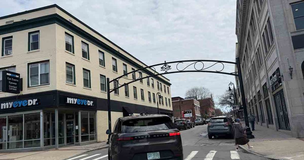 A street scene with cars, pedestrians, and buildings, including a "myeyedr." eye care store. An arched metal sign crosses the street, and the sky is cloudy.
