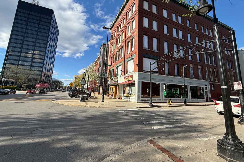 A sunny city street corner with a red brick building, storefronts, parked cars, and a modern glass office tower in the background. The sky is partly cloudy with patches of blue.