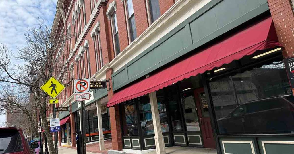 A row of brick buildings with green and red awnings over storefronts, a yellow pedestrian crossing sign, and a no-parking sign on a city sidewalk with bare trees.