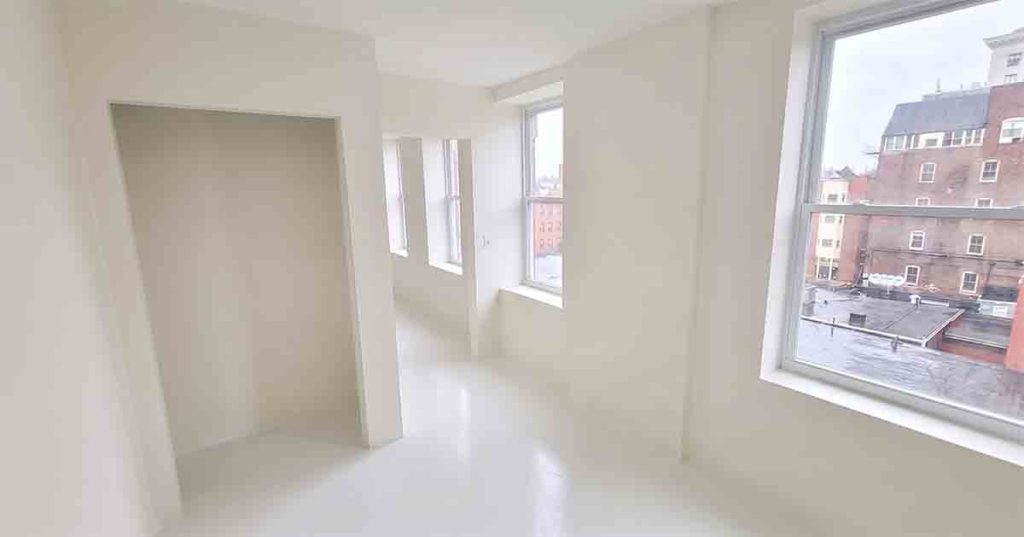 A bright, empty room with white walls and floors, featuring large windows that let in natural light and provide a view of surrounding buildings.