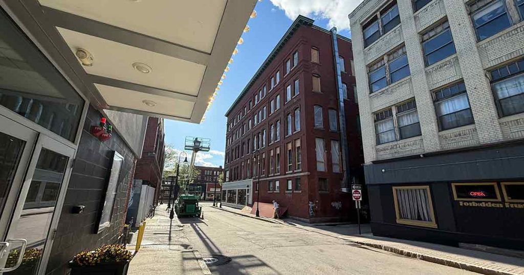 A narrow city street with brick and white buildings, a green truck parked on the left, and a restaurant with a neon "OPEN" sign on the right under a sunny blue sky.