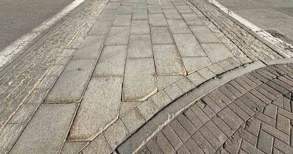 A close-up view of a sidewalk and street corner, showing different textures and patterns of paving stones and bricks, with a curb separating the sidewalk from the street.