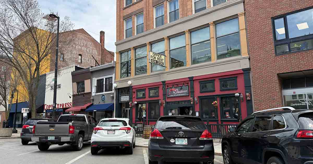 Several cars are parked on a city street in front of restaurants and shops. A large "For Rent" sign is visible in the second-floor window of a brick building. Trees line the sidewalk.