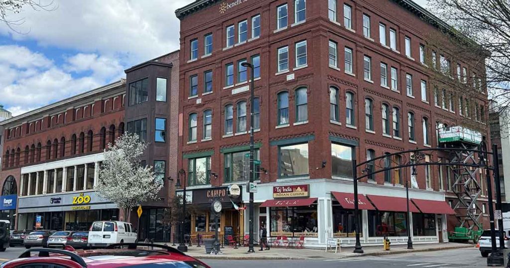 A street corner with historic red-brick buildings, storefronts, large windows, and awnings. Cars are parked along the street, and a white-blossomed tree stands near the sidewalk under a partly cloudy sky.