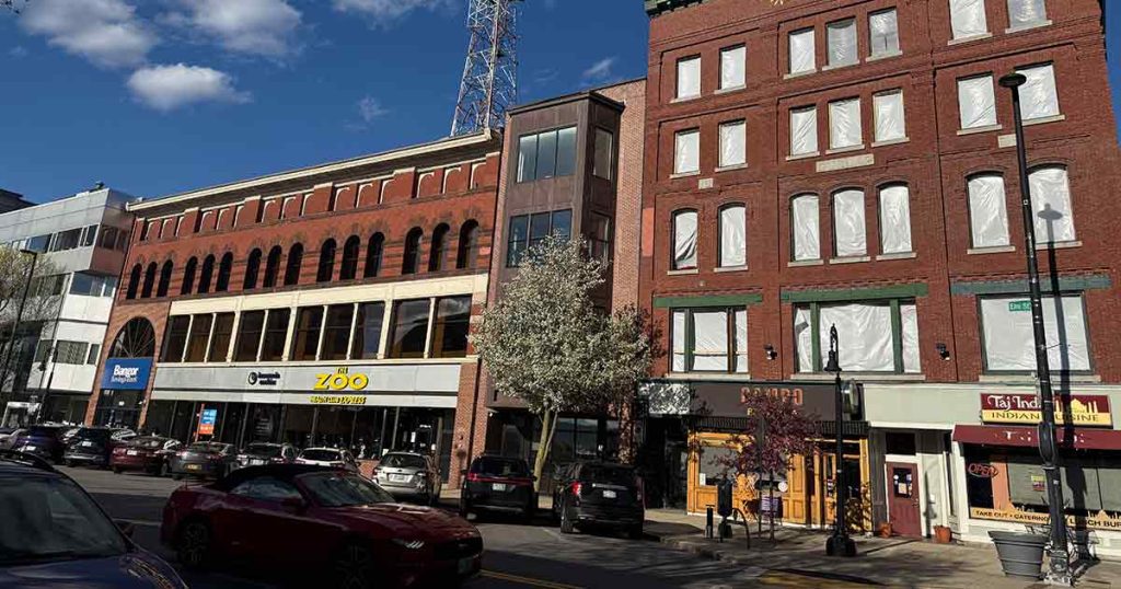 A sunny street scene with red brick buildings, shops on the ground floor, parked cars, flowering trees, and a blue sky with a few clouds overhead. Some windows on upper floors are covered with white sheets.