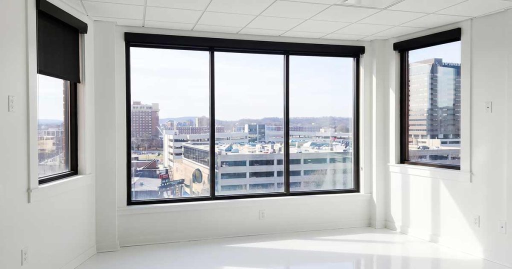 A bright, empty office room with large windows offering a cityscape view of buildings and a clear sky. The room has white walls, a white floor, and black window shades partially rolled up.
