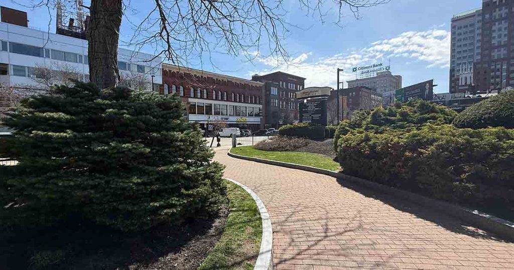 A curved brick pathway winds through a park with green bushes and trees, leading toward buildings and shops under a sunny blue sky in an urban area.