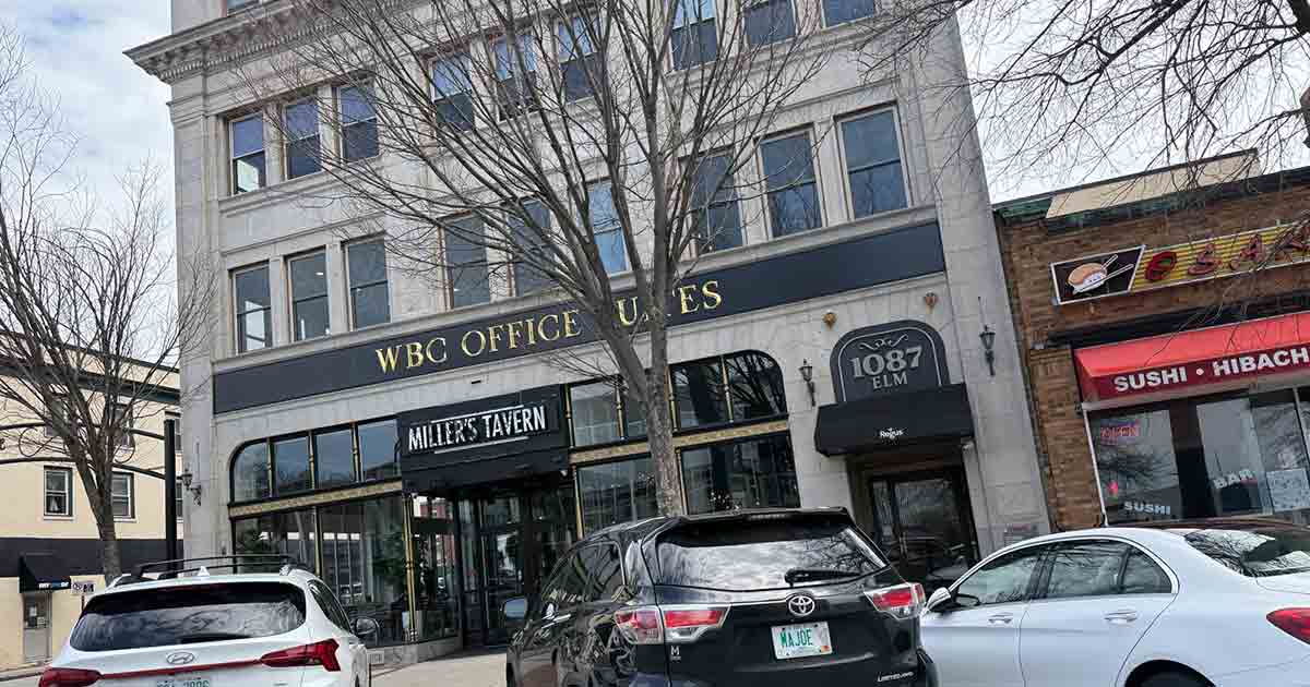 Three cars are parked in front of a multi-story building with signs for "WBC Office Suites," "Miller's Tavern," and "1087 Elm." Leafless trees line the sidewalk under a cloudy sky.
