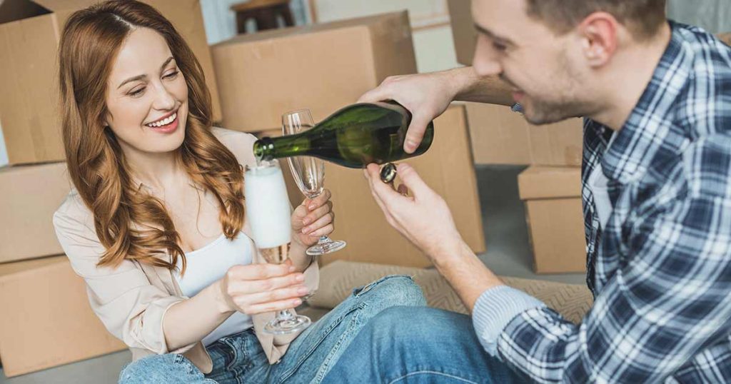 A smiling woman holds a champagne glass while a man pours champagne. They are sitting on the floor surrounded by cardboard moving boxes.