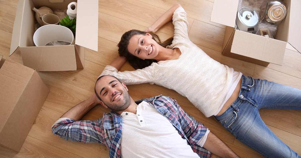 A smiling couple lies on the wooden floor among open cardboard boxes, appearing relaxed and happy, possibly after moving into a new home.