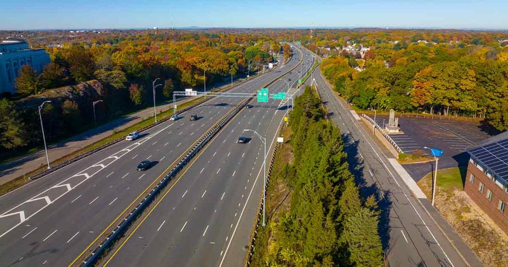 Aerial view of a wide highway with light traffic, surrounded by trees with autumn foliage, buildings, and clear blue sky in the background.