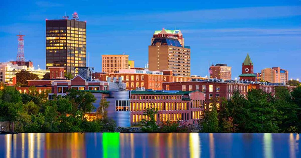City skyline at dusk with modern and historic buildings, trees in the foreground, and water reflecting colorful lights from the buildings.