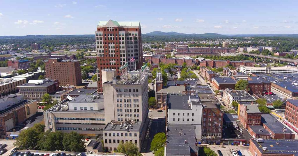 Aerial view of a city with tall buildings, mid-rise structures, and distant green hills under a clear blue sky. Trees and cars are visible along the streets.