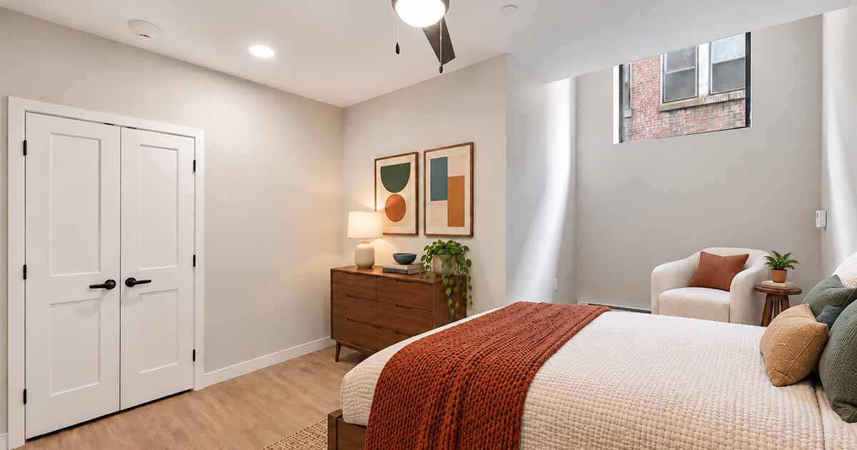 A bedroom with a bed topped with a textured rust-colored throw, a wooden dresser with two framed geometric prints above it, a white accent chair, and a high window.