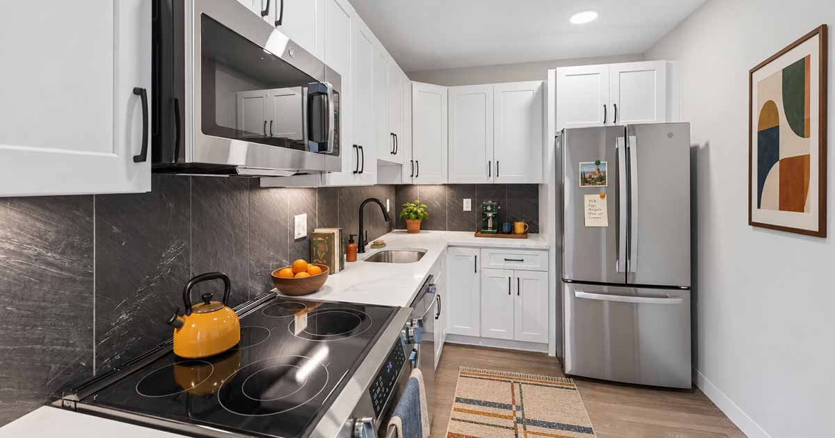 A modern galley kitchen with white cabinets, stainless steel appliances, a black electric stovetop, and dark stone backsplash.