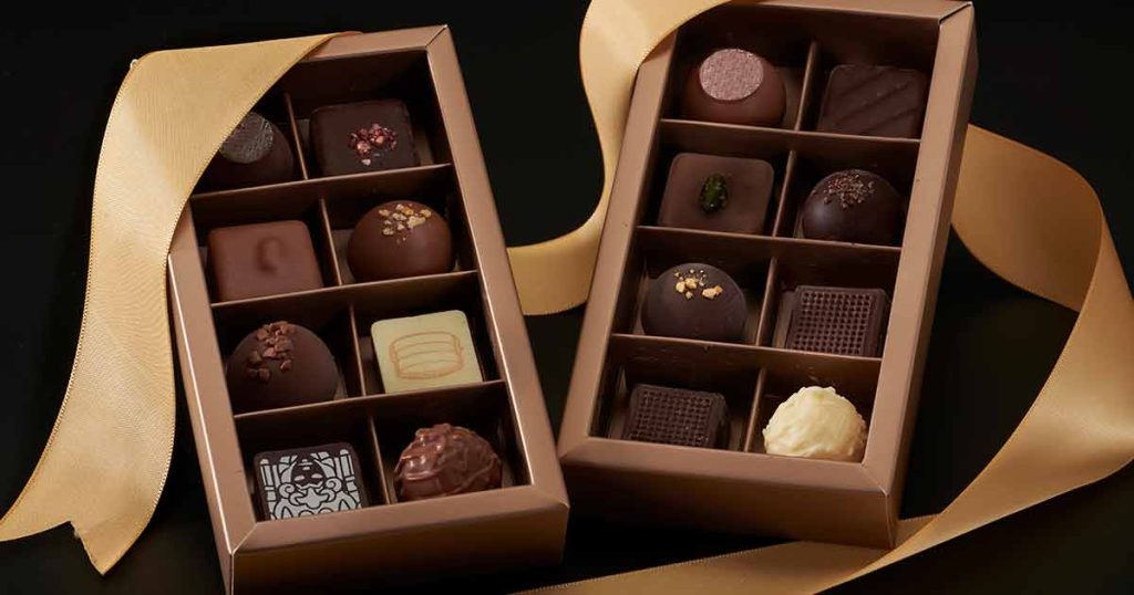 Two rectangular gift boxes filled with an assortment of assorted gourmet chocolates, decorated with a gold ribbon.