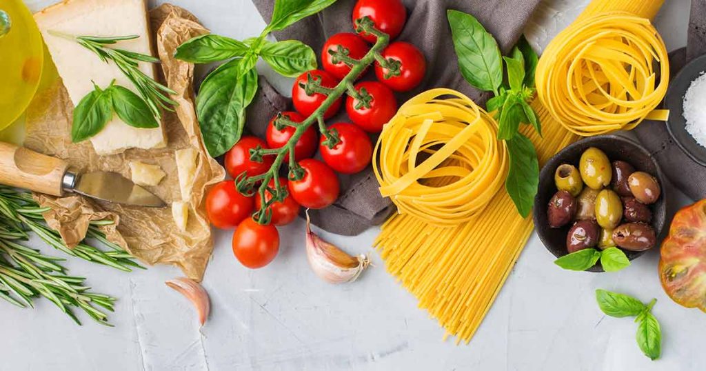 Ingredients for pasta including nests of fettuccine, a vine of cherry tomatoes, basil, a block of parmesan, olives, rosemary, and garlic cloves.