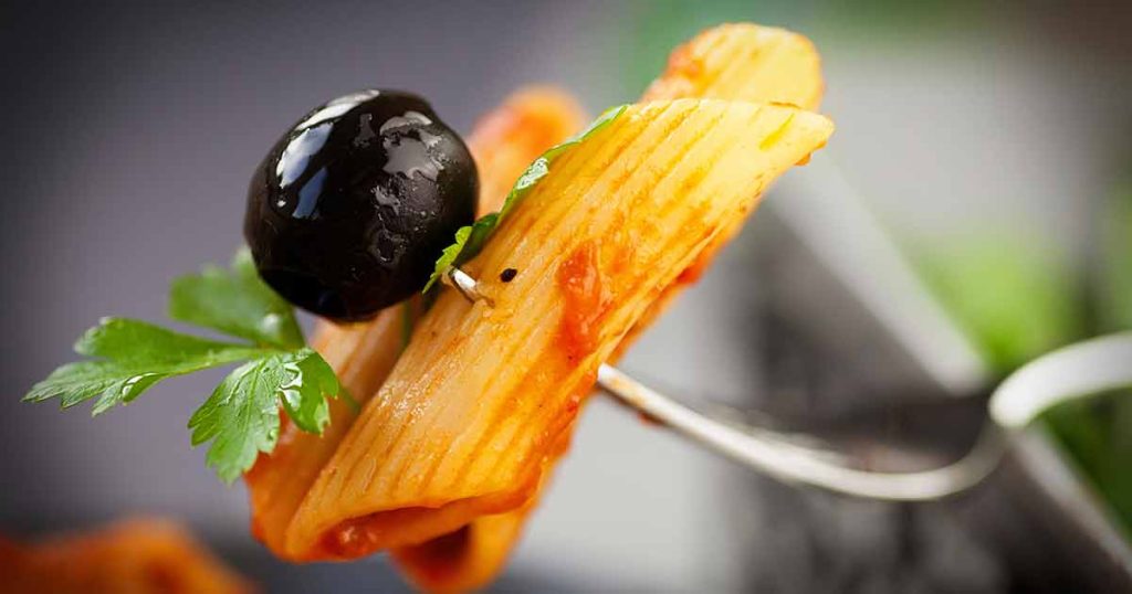 A fork holding a piece of penne pasta with tomato sauce, a black olive, and a parsley leaf.