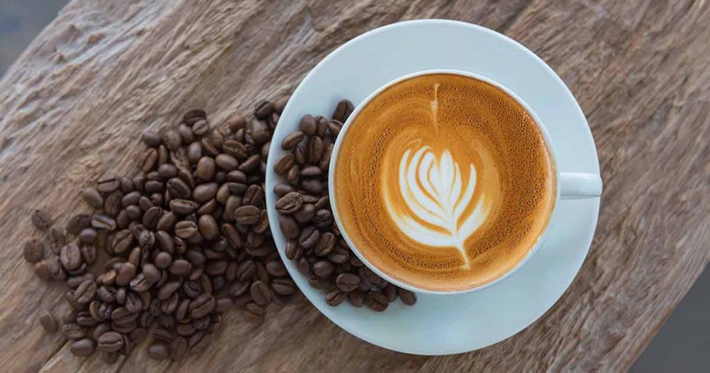 A cup of latte art coffee on a saucer, surrounded by a pile of roasted coffee beans on a wooden surface.